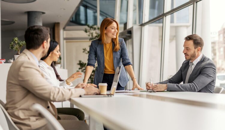 3 formally dressed people sat around a large table having a meeting, with a red-headed woman stood at the head of the table laughing