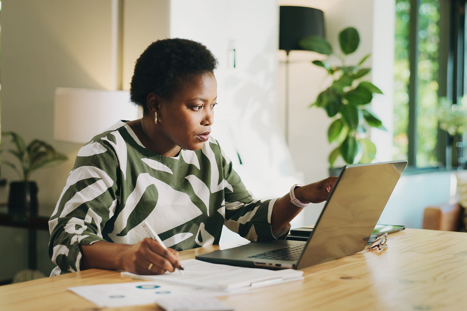 woman in green top looking at laptop and writing down financial details onto paper